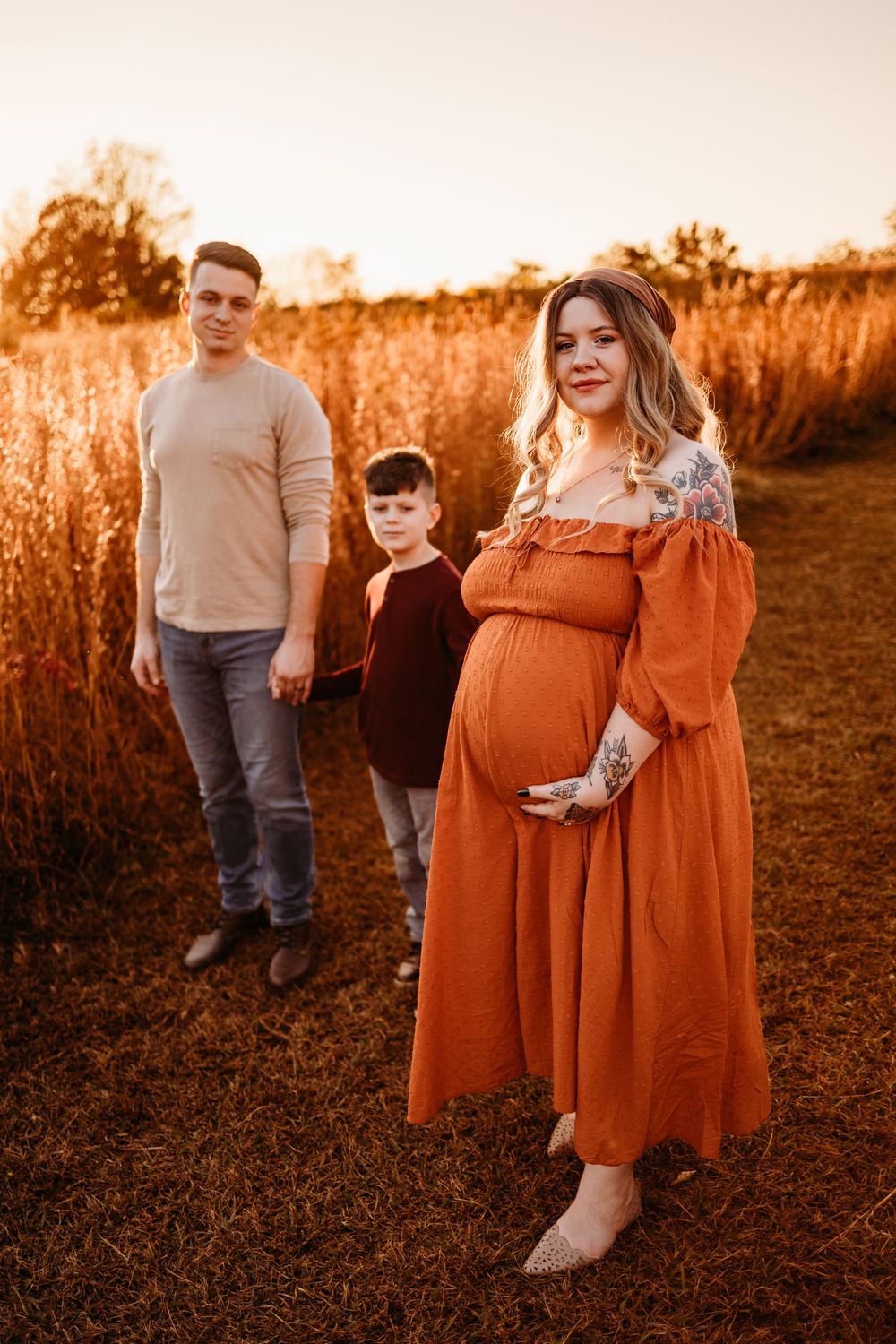 Pregnant woman in orange dress with husband and son at golden hour in a field.