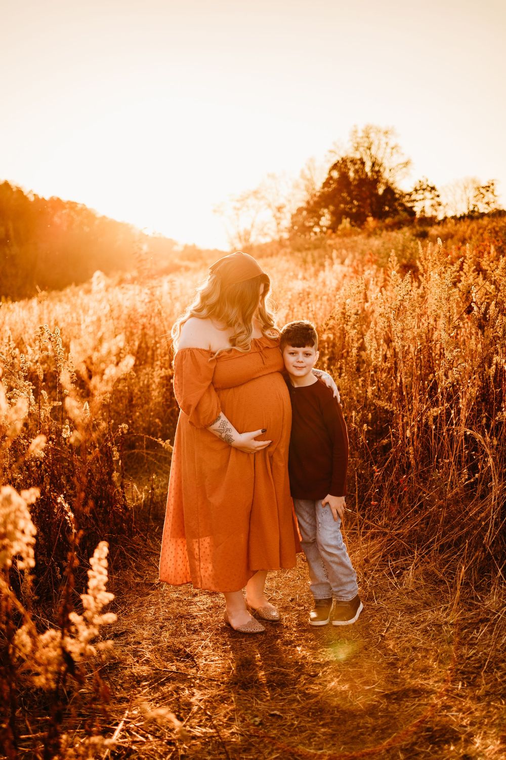Pregnant woman in rust dress with young son hugging her belly at golden hour in autumn field.