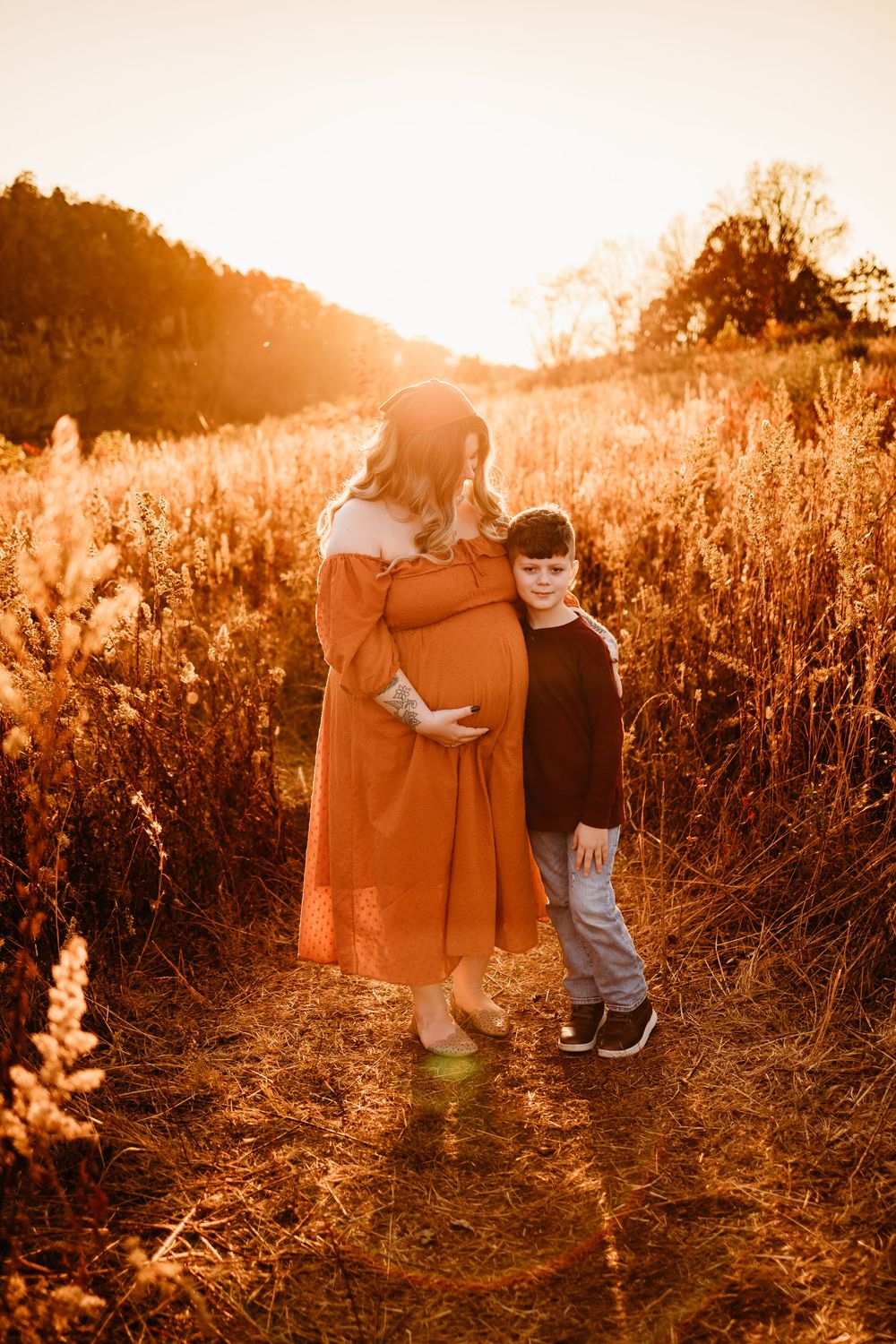 Pregnant woman in orange dress with young boy in golden sunset field during maternity photo session.