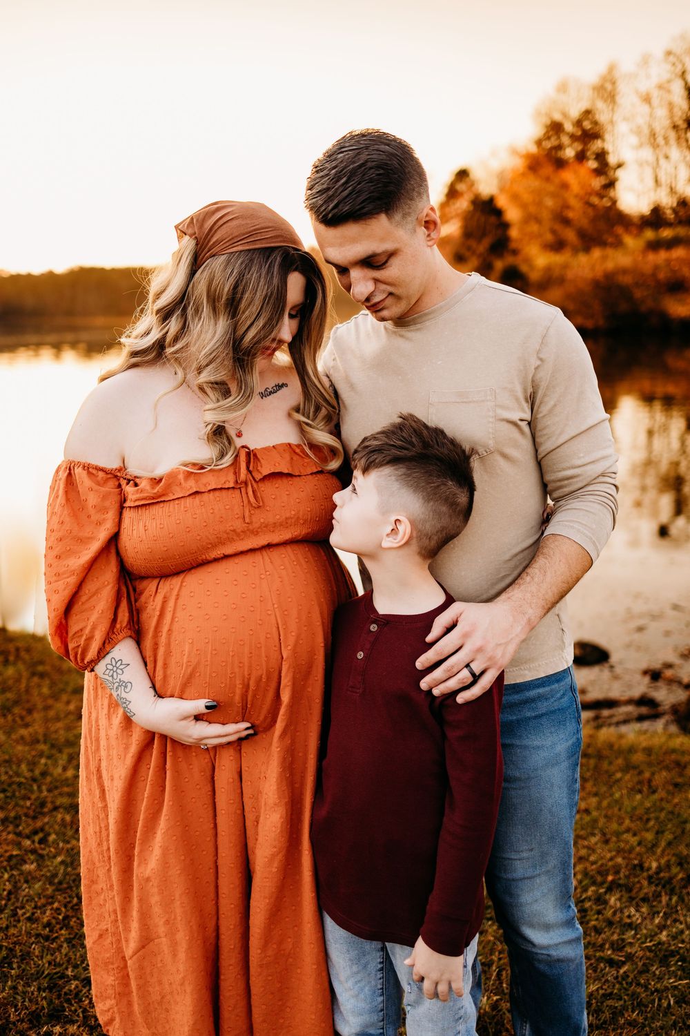Family maternity photo at golden hour by a lake, pregnant mom in orange dress with partner and son.
