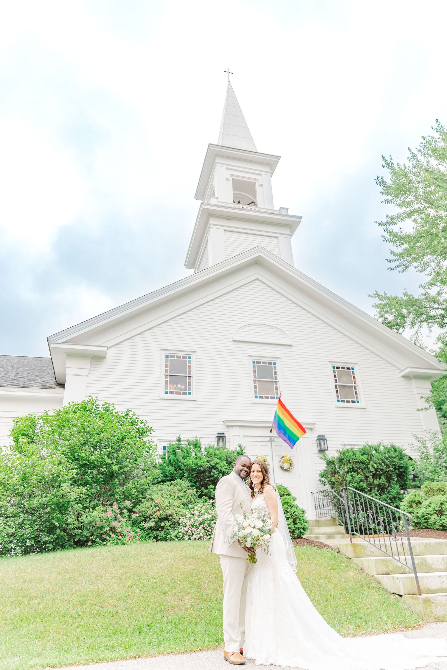 White church with a steeple and pride flag displayed in front on a sunny summer day.