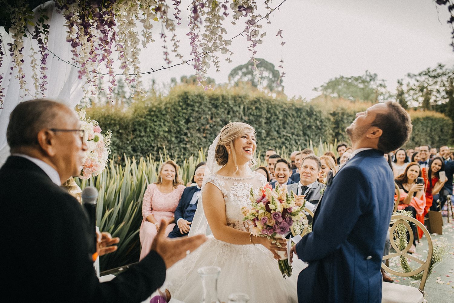 Bride laughing joyfully at outdoor wedding ceremony with colorful bouquet under wisteria.