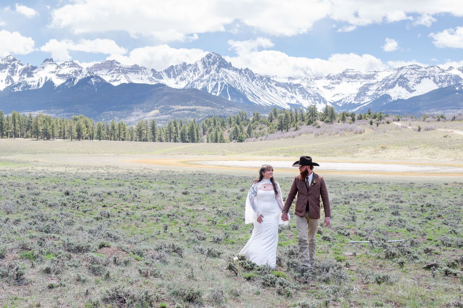 Bride and groom holding hands in mountain meadow with snow-capped peaks, rustic western wedding attire, Idaho wilderness.
