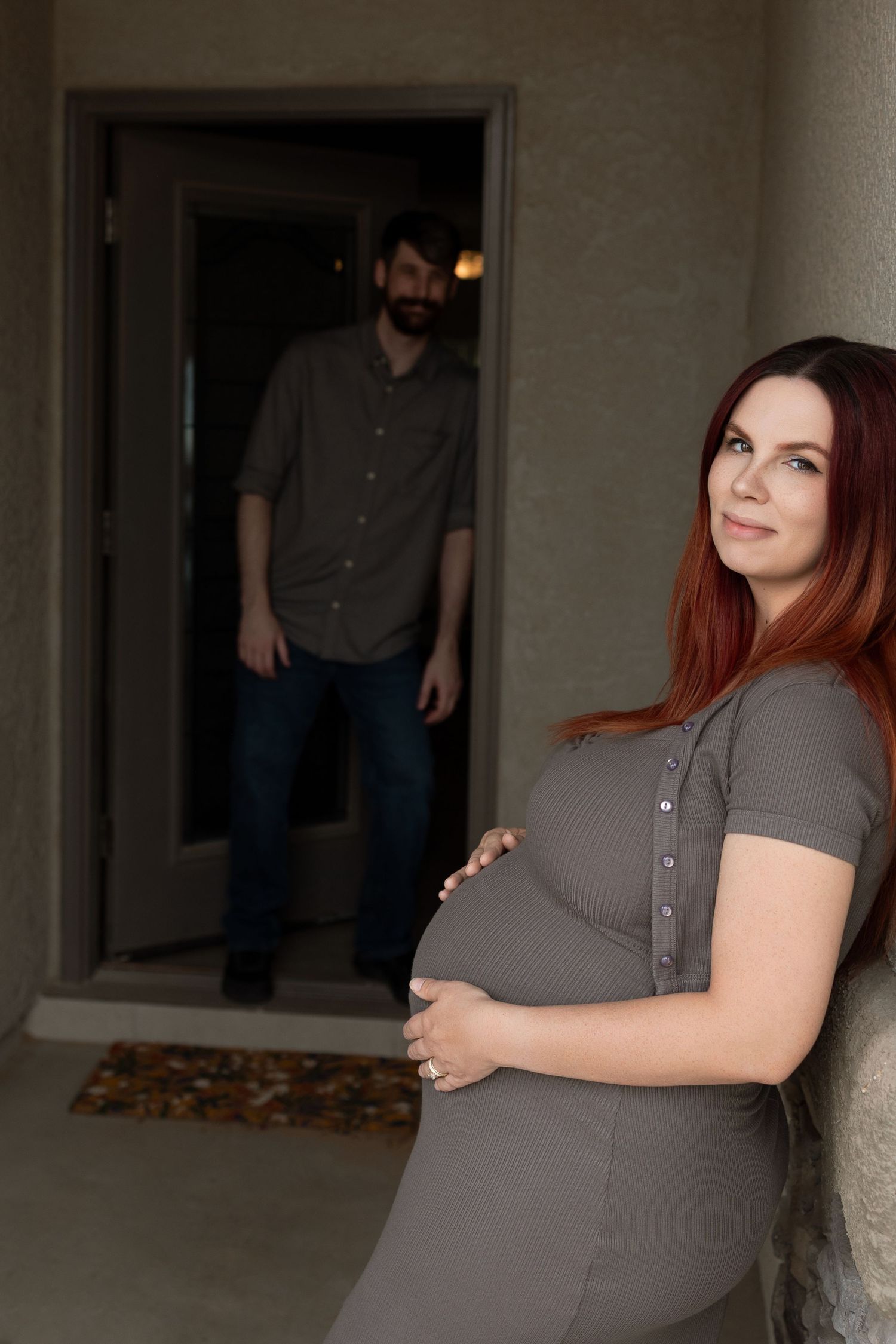 Pregnant woman in gray dress cradling baby bump outdoors while man stands in doorway behind her.