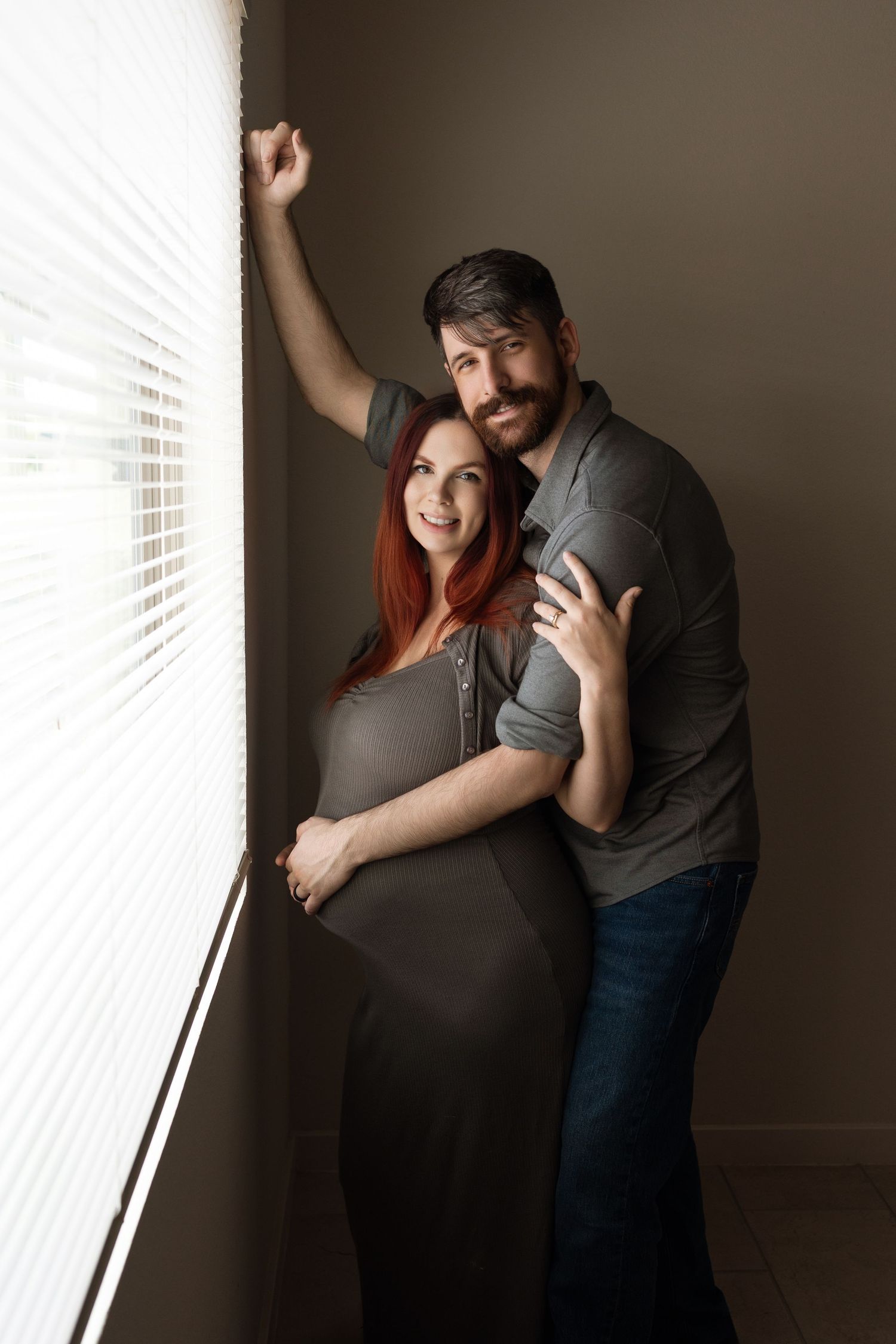 Pregnant woman in gray dress embraced by man near window blinds, maternity couple portrait.
