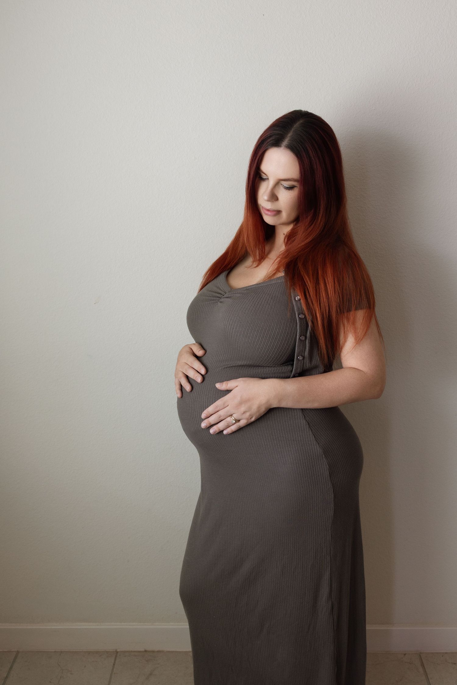 Pregnant woman in olive maxi dress gently cradling baby bump against light wall.