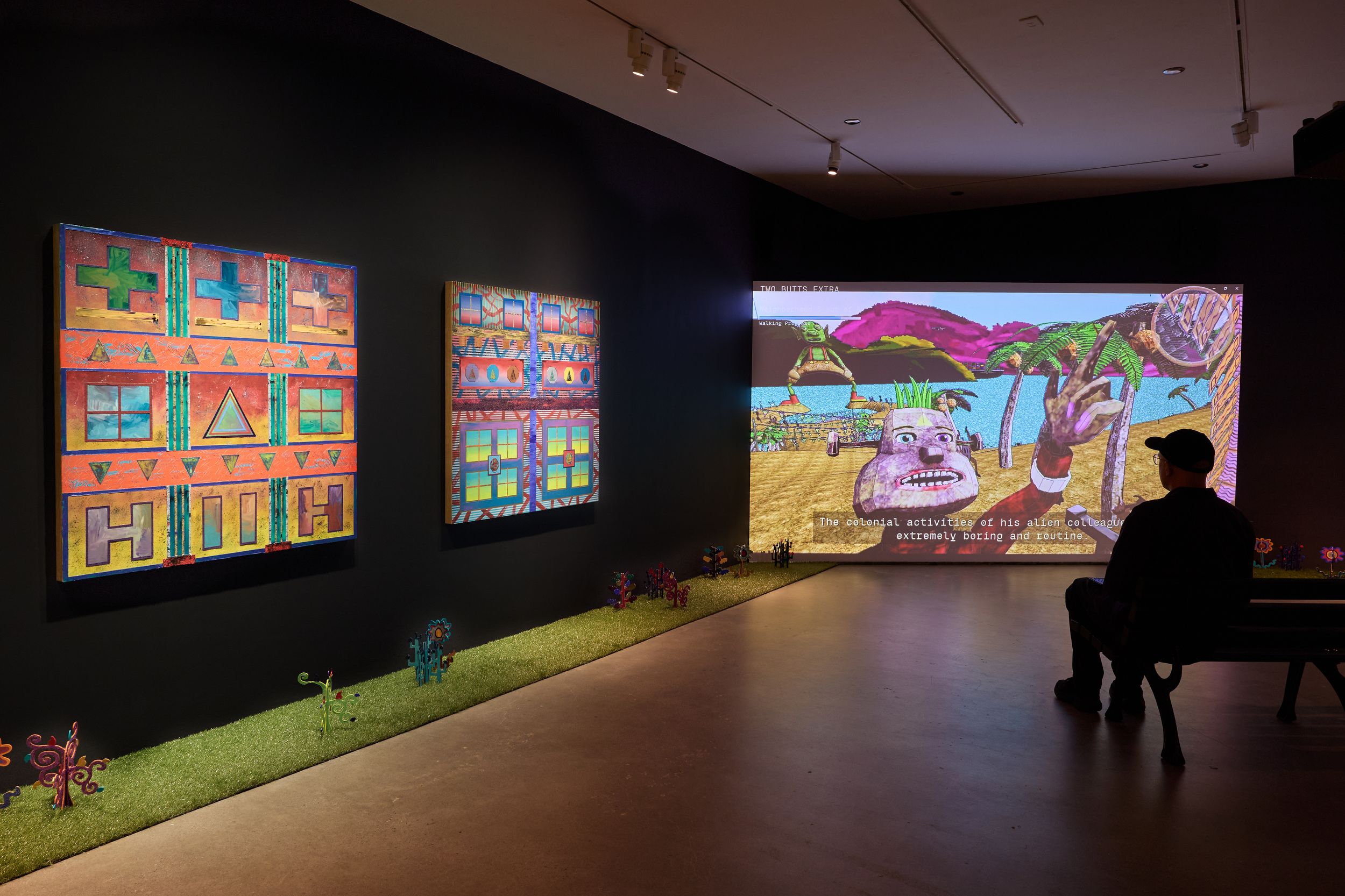 Art gallery display with colorful paintings and a large video projection, visitor silhouetted viewing animated scene.