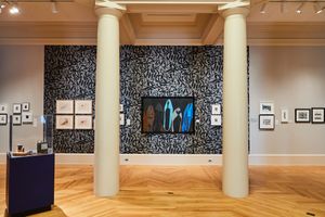 Art gallery interior with patterned blue wall, framed artwork, white columns, and herringbone wood floors.