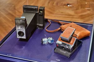 Two vintage Polaroid cameras displayed on a blue velvet surface, with a brown leather strap camera case.