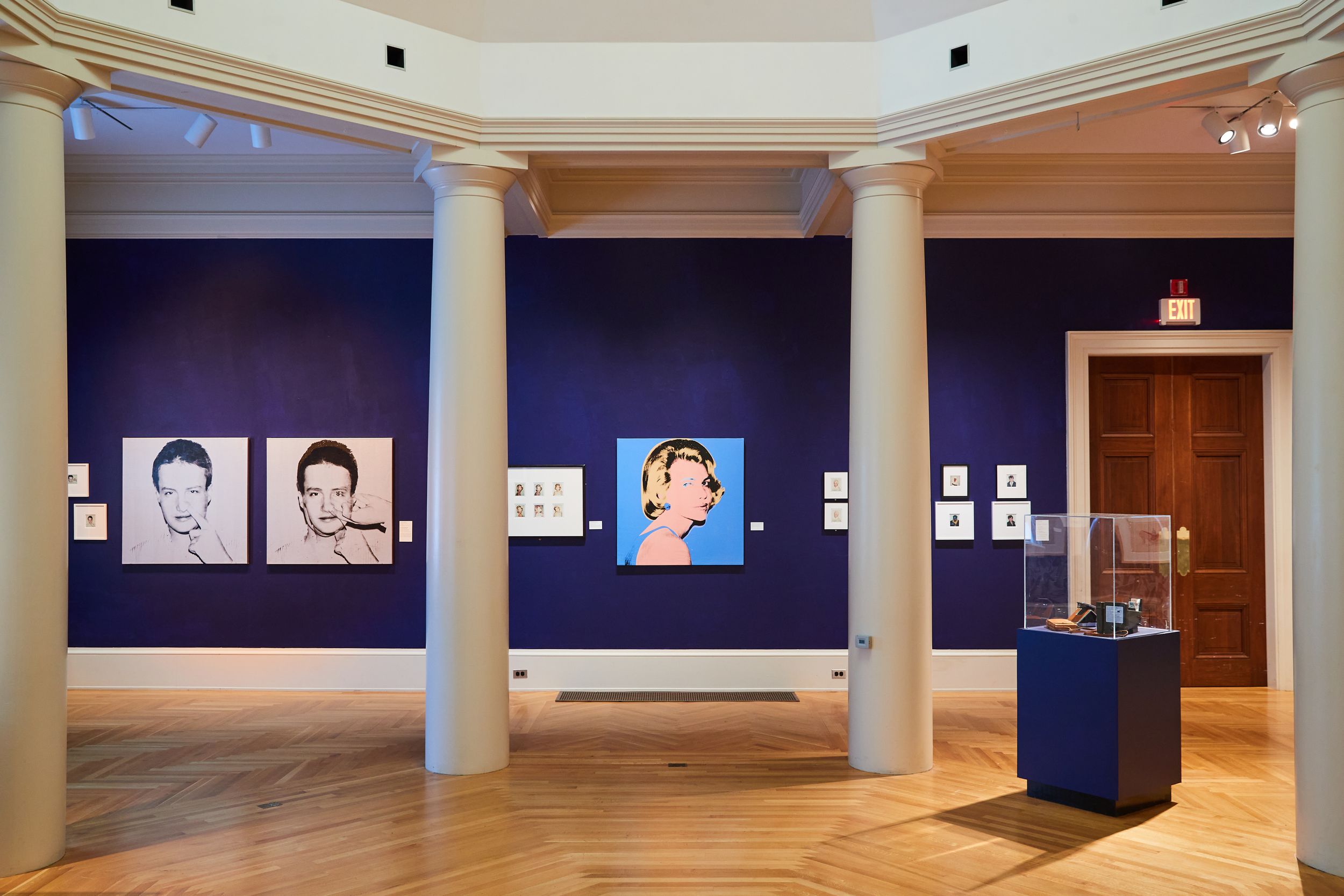 Gallery with navy blue walls displaying pop art portraits and white columns on a hardwood floor.