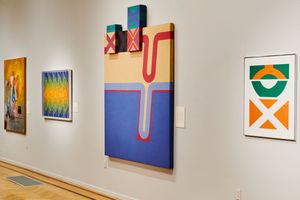 Art gallery wall displaying colorful abstract paintings including geometric shapes in blue, red, green, and yellow on white walls.