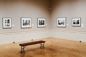 Museum gallery with black-and-white framed photographs on gray walls, a wooden bench on herringbone parquet flooring.