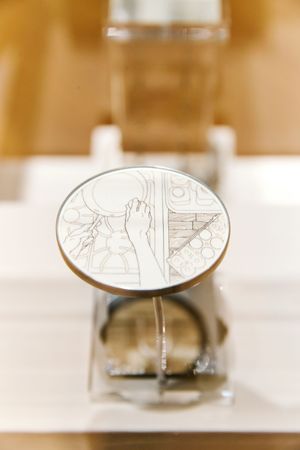 Oval decorative coin with engraved hand artwork displayed on a stand inside a glass jar exhibit.