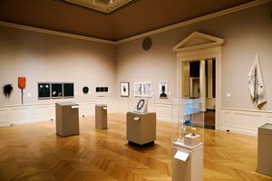 Elegant art museum gallery with parquet floors, pedestals displaying sculptures, and framed artwork on taupe walls.