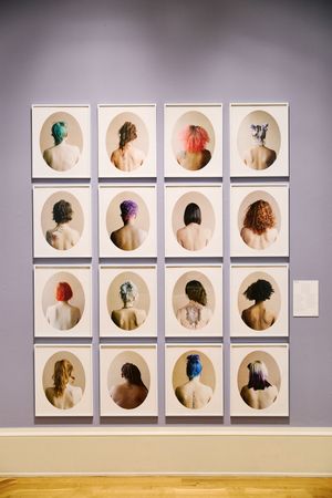 Gallery wall displaying 16 framed oval portraits of figures seen from behind, featuring diverse colorful hairstyles on a lavender wall.