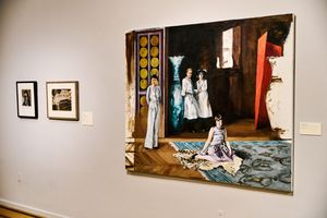 Art gallery wall displaying a large figurative painting of women in white dresses alongside smaller framed black and white photographs.