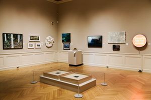 Art gallery interior with herringbone wood floors, beige walls displaying paintings, sculptures, and mixed media exhibits.