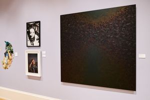 Gallery wall displaying mixed artworks including a large dark textured painting, black and white photos, and colorful sculpture.