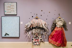 Gallery wall displaying two framed artworks and two ornate traditional garments on hangers, featuring fringed and embroidered designs.