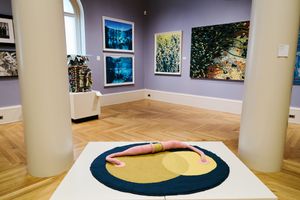 Art gallery interior featuring a sculptural figure on a circular rug, surrounded by colorful paintings on lavender walls.