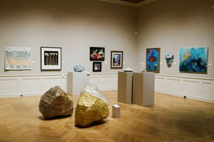 Art gallery interior with gold sculptural rocks, pedestals, and colorful paintings displayed on beige walls with parquet flooring.