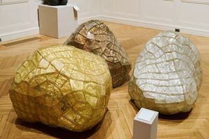 Three large sculptural boulder-like artworks with cracked golden and brown textures displayed on a parquet floor in a gallery.