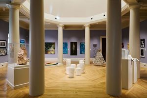 Elegant art museum gallery with white columns, herringbone wood floors, and colorful paintings and sculptures on display.