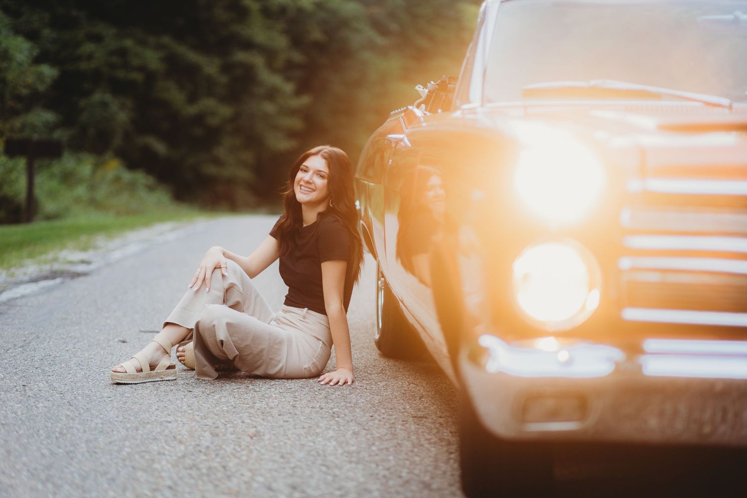 Young woman in casual outfit sitting on road leaning against vintage black car with glowing headlight. stoney creek senior session