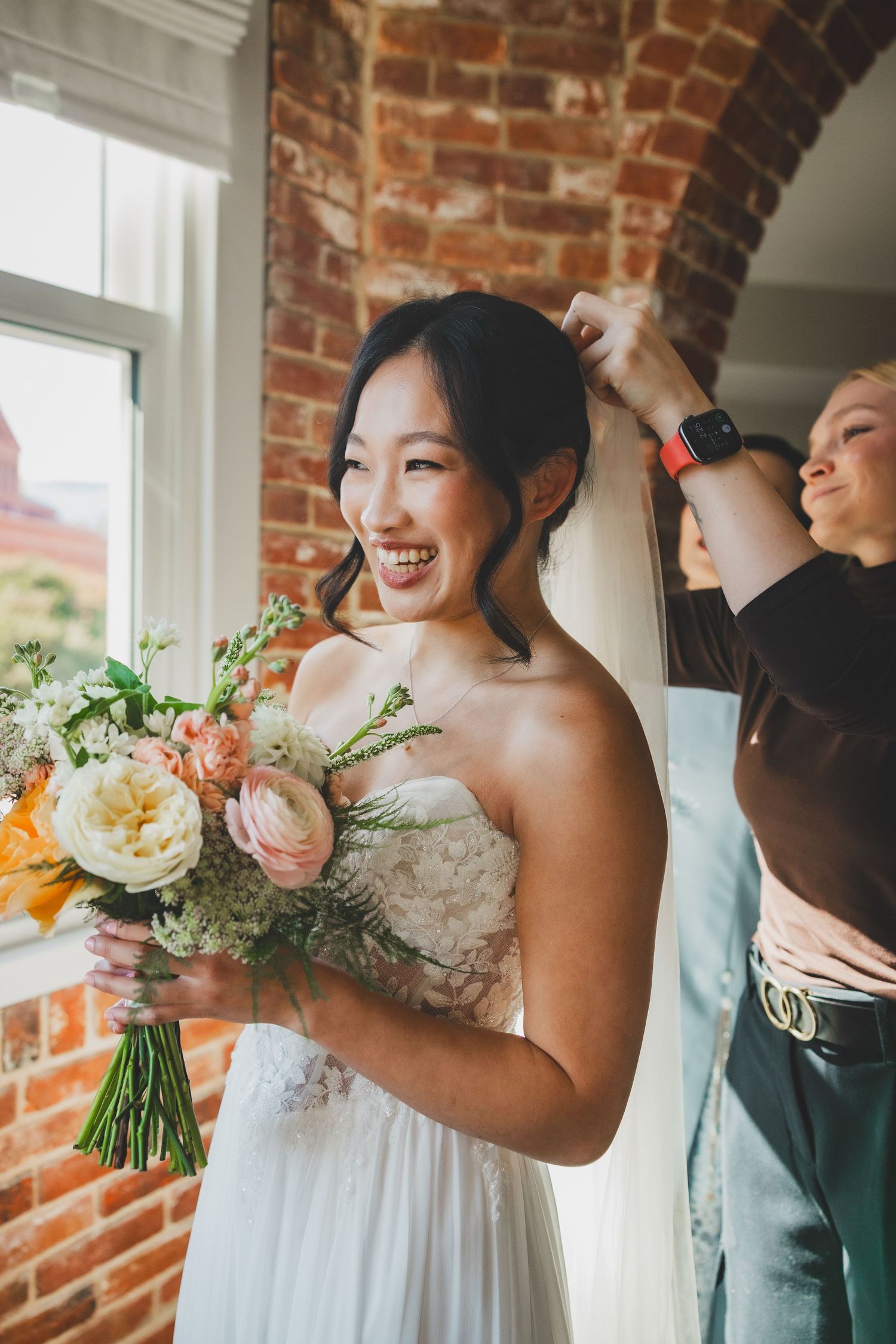 Smiling bride in strapless white gown holds floral bouquet while assistant adjusts veil near brick wall window.