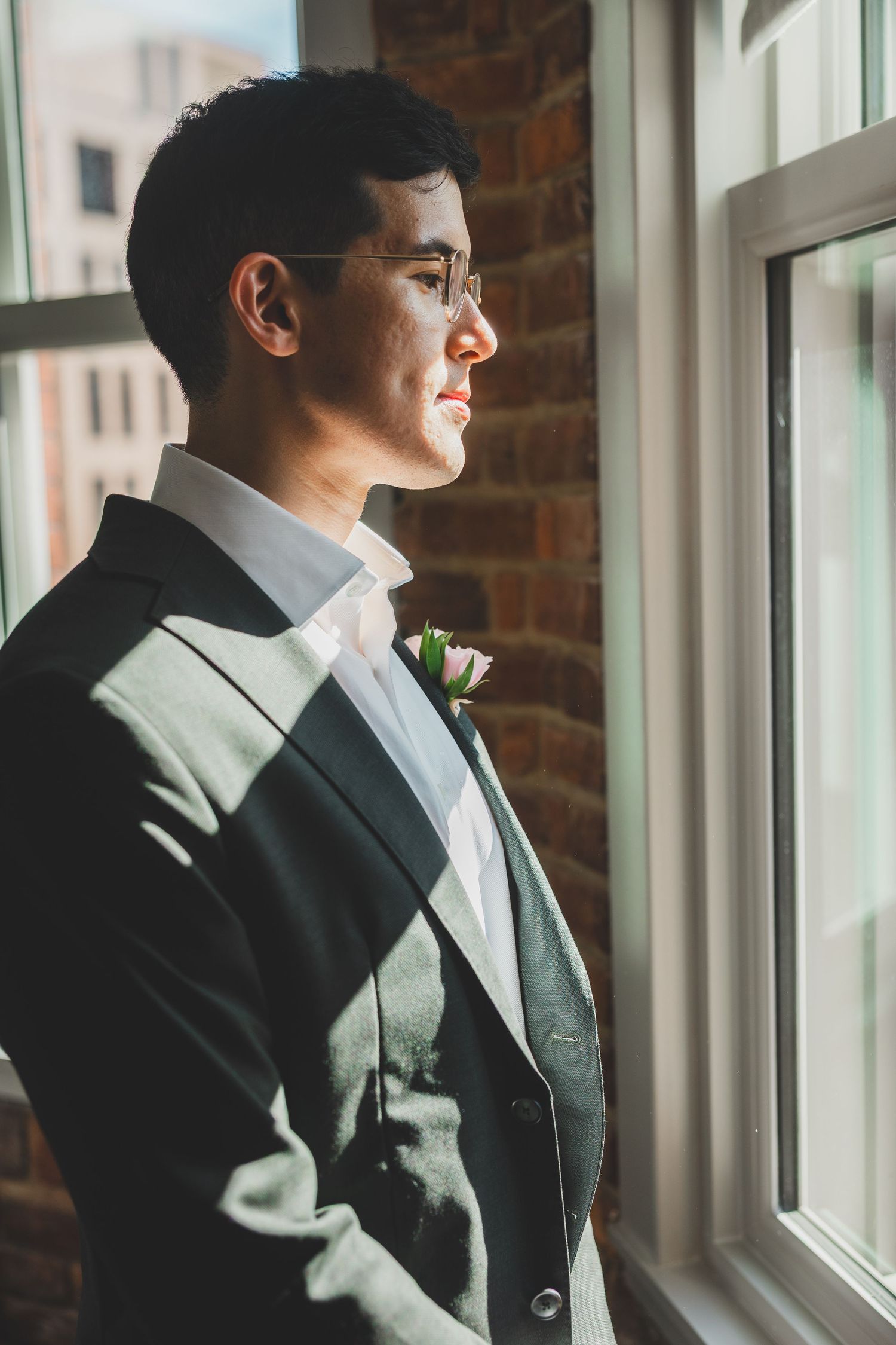 A groom in a gray suit with a boutonniere gazes thoughtfully out a sunlit window against a brick wall.