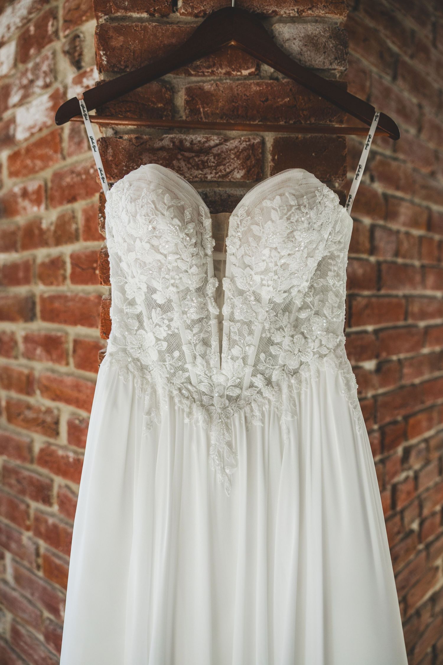 Elegant white lace bodice wedding dress hanging on wooden hanger against rustic brick wall.