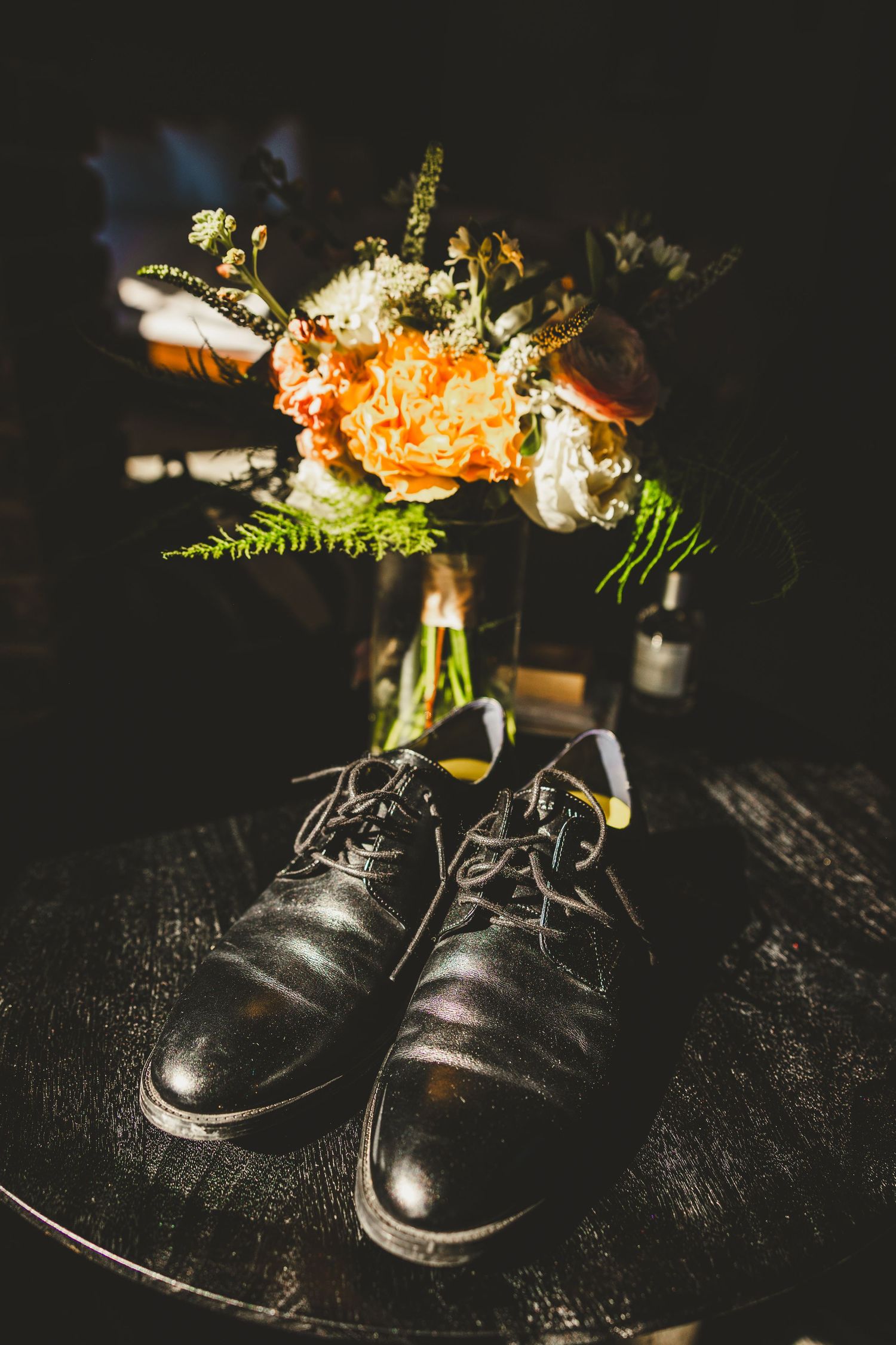 Black leather lace-up dress shoes beside a vibrant orange and white wedding bouquet on a dark wooden table.