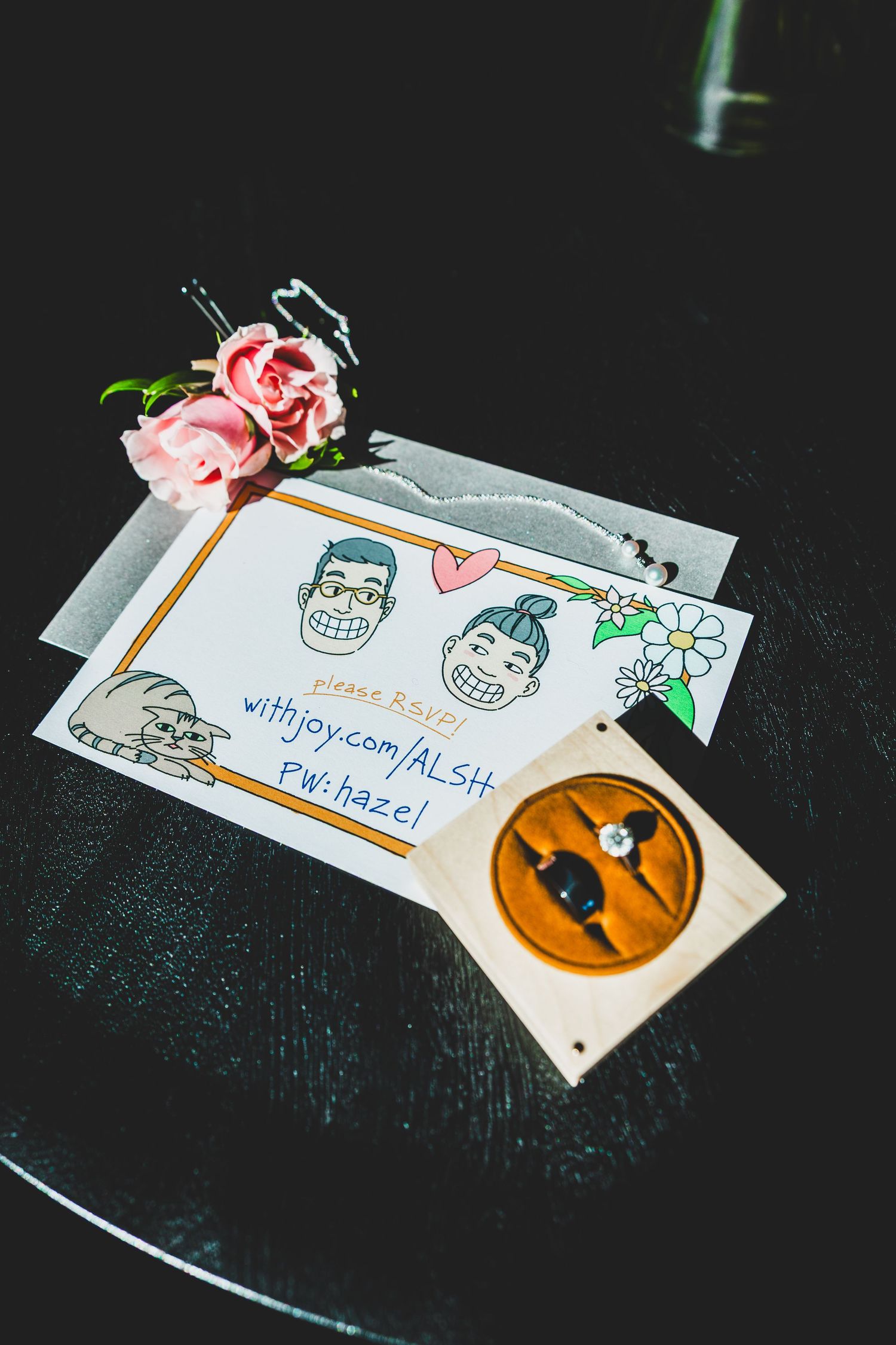 Wedding ring box and illustrated couple card with pink roses on dark table, featuring withjoy.com RSVP details.