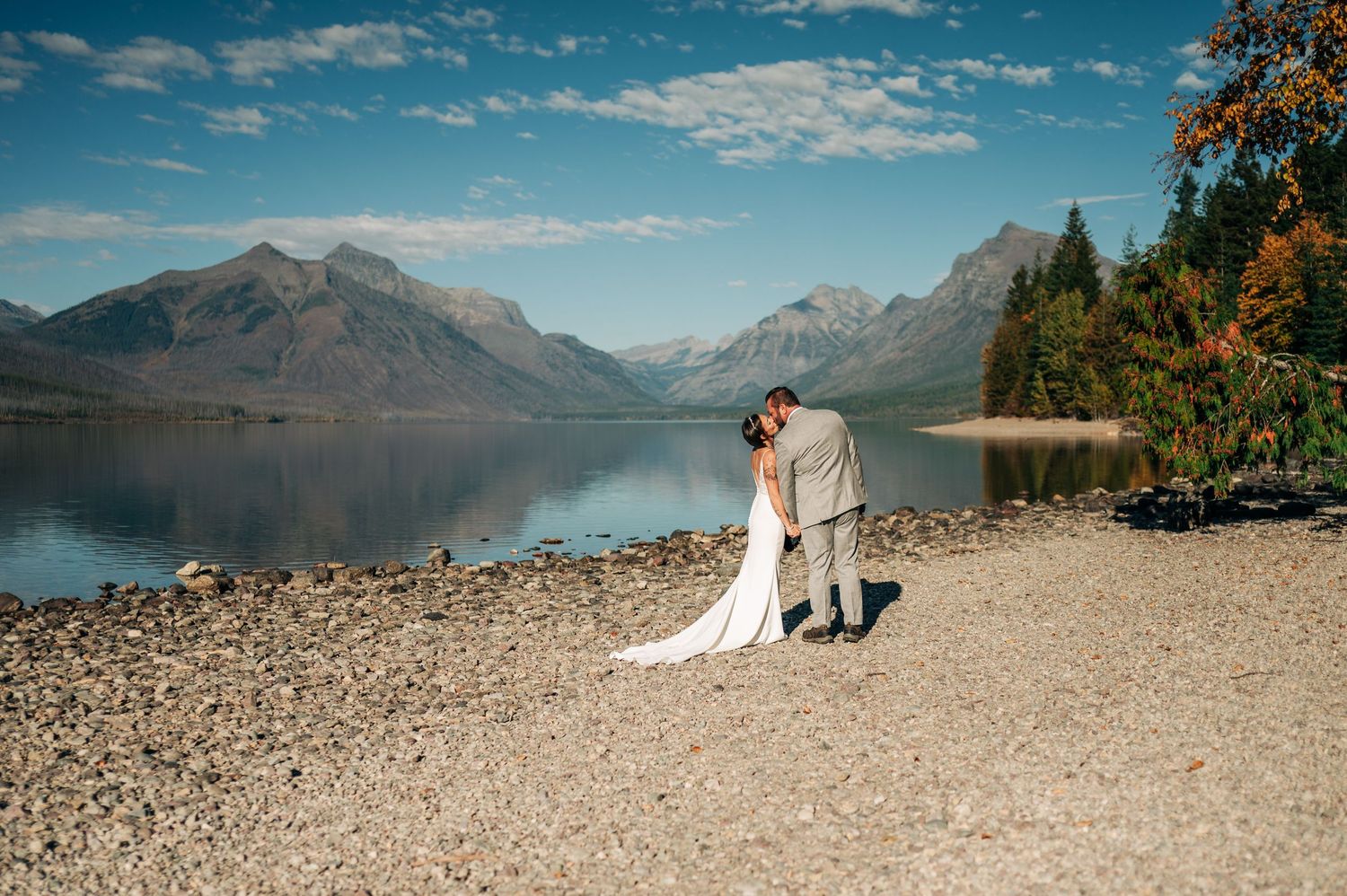 Bride and groom kissing on rocky lakeshore with stunning mountain backdrop under blue sky.