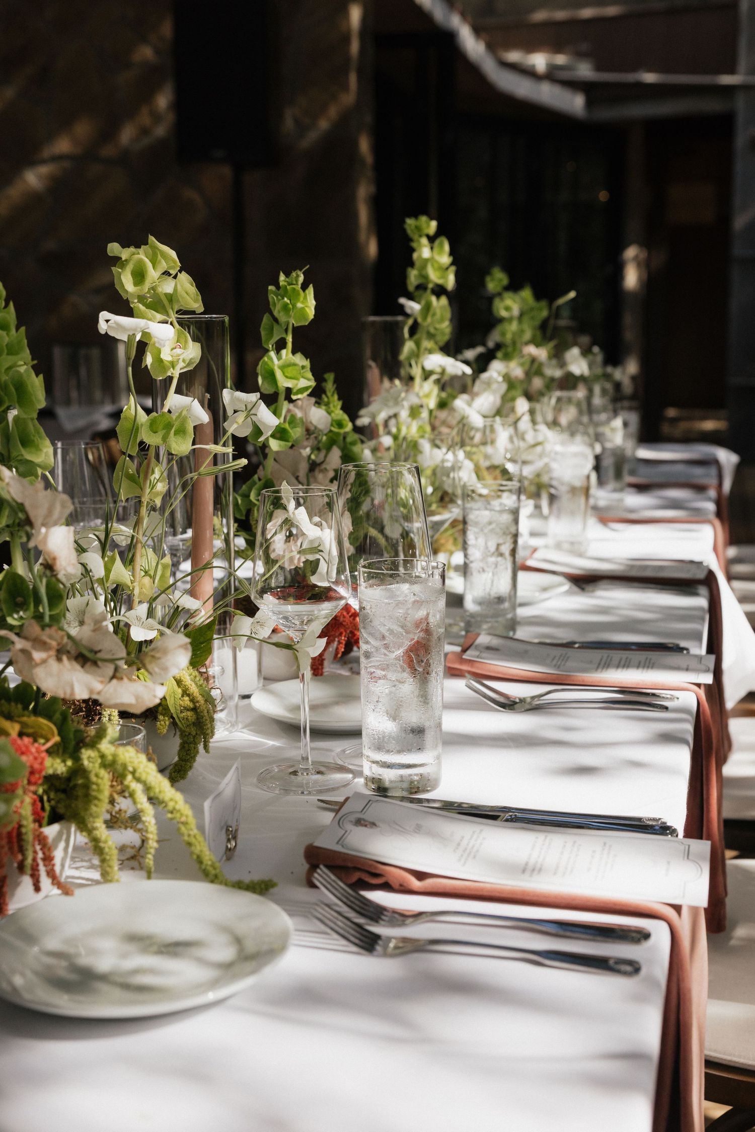 Elegant wedding reception table with white linen, copper cutlery, glass vases, and lush green floral centerpieces.