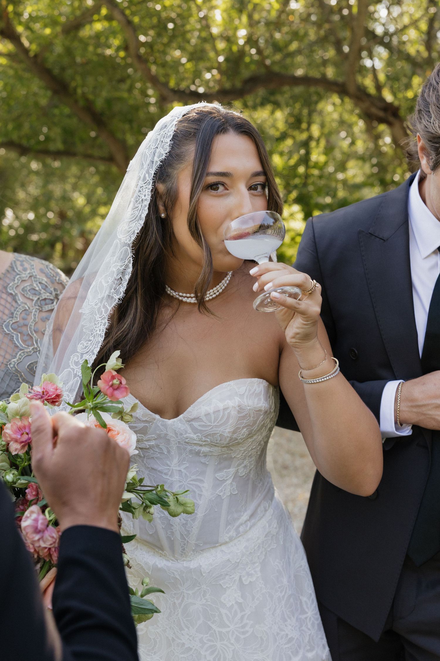 Bride in lace wedding gown and veil sips from wine glass outdoors, holding pink bouquet beside groom in black suit.