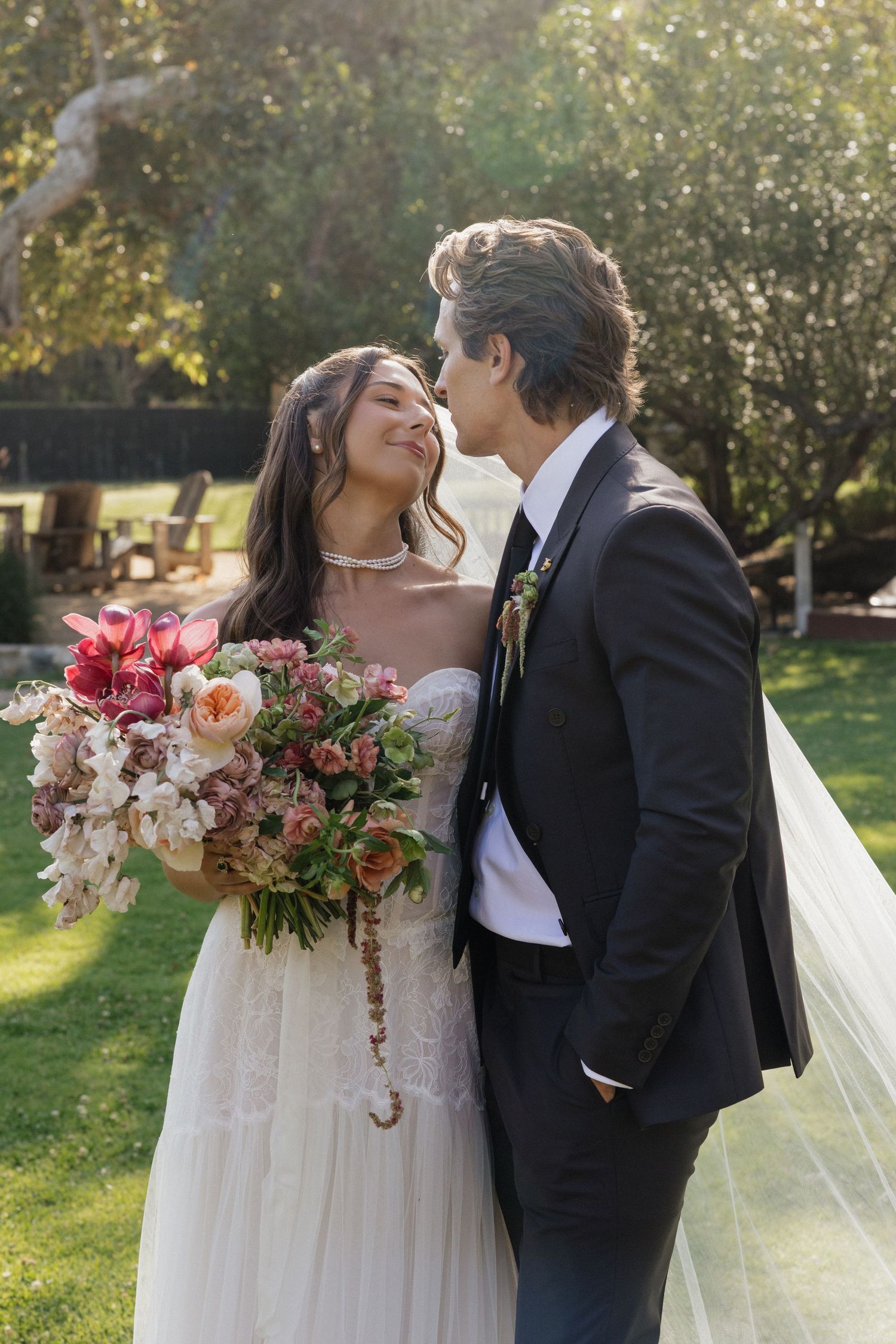 Bride and groom share a romantic moment outdoors, bride holding a lush pink and white bouquet with veil flowing.