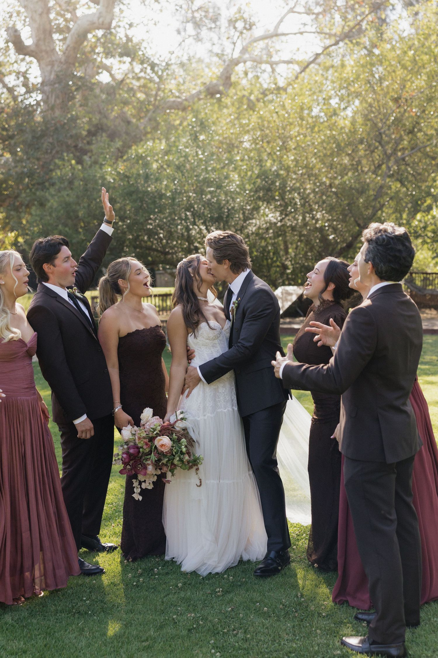 Wedding party celebrates as bride and groom share a kiss outdoors, surrounded by bridesmaids in burgundy gowns and groomsmen.