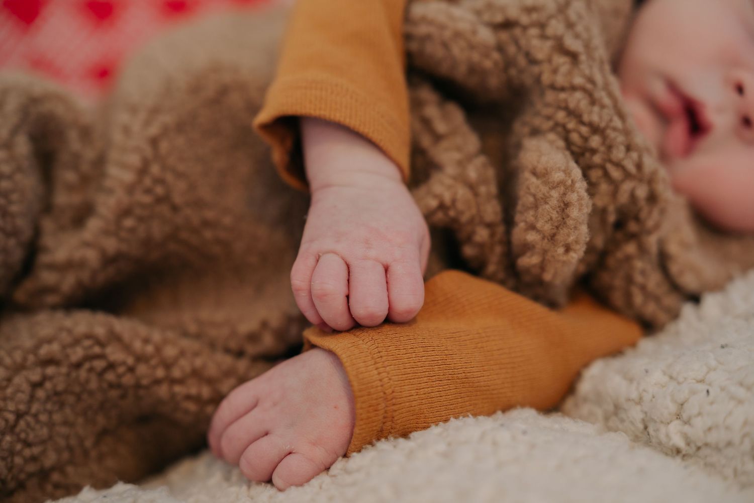 Close-up of a sleeping baby's tiny hands and feet in a cozy brown teddy bear outfit on a soft blanket.