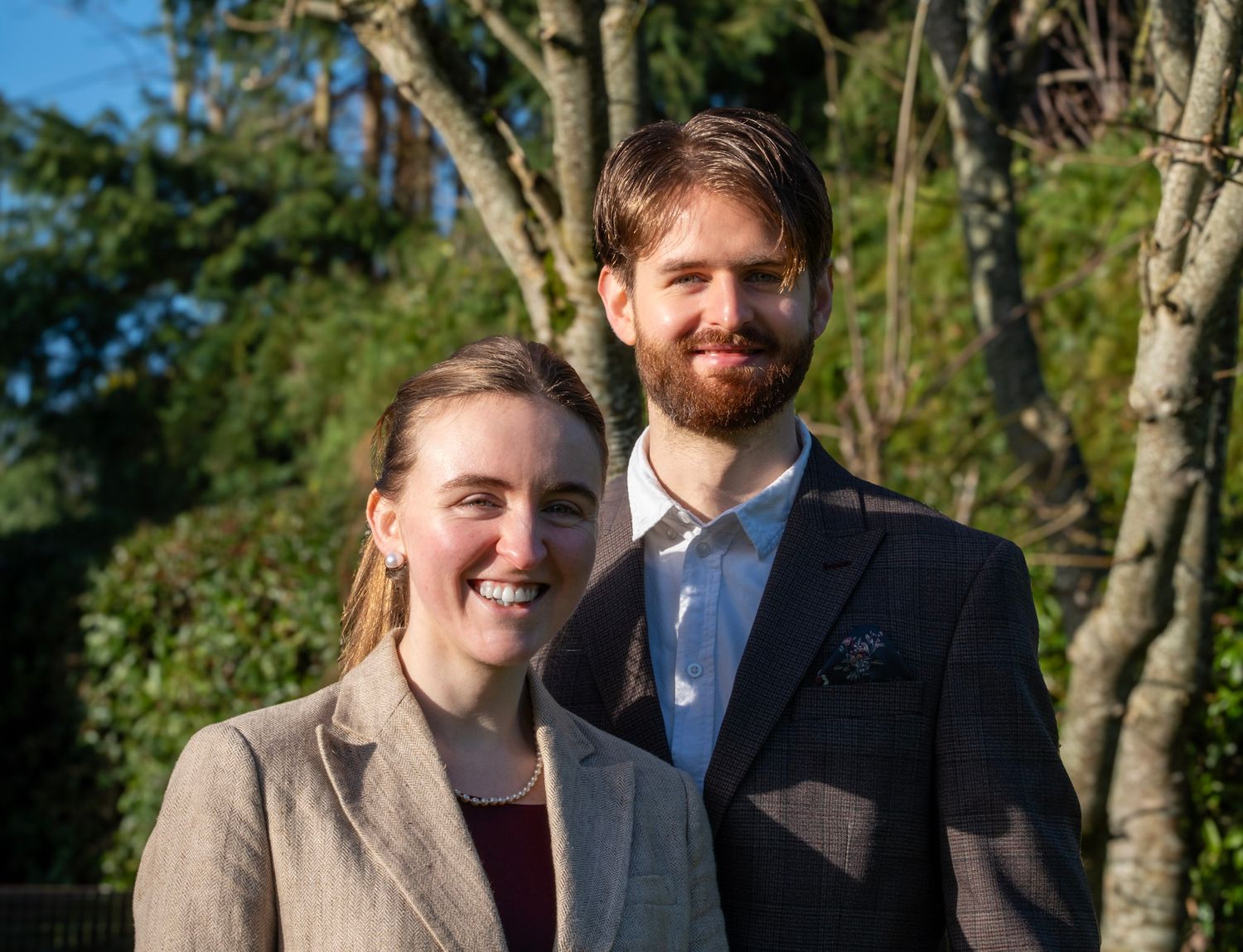 Smiling couple posing outdoors in formal attire with lush green trees in the background.