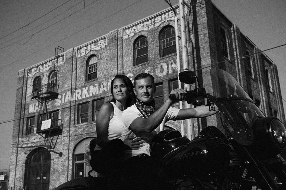 Black and white photo of a couple posing on a motorcycle in front of a vintage brick building with faded signage.