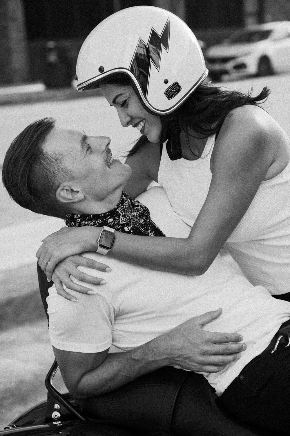 Black and white photo of a romantic couple embracing on a motorcycle, woman wearing a white helmet smiling at man.