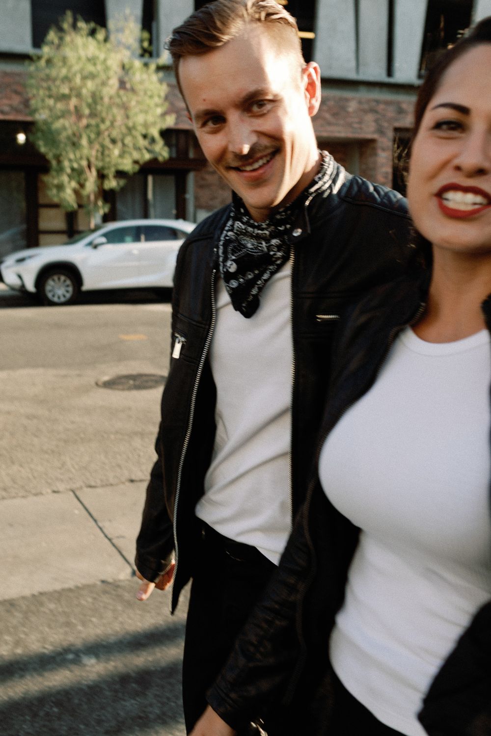 A smiling man in a black leather jacket and bandana walks with a woman in a white top on a city street.