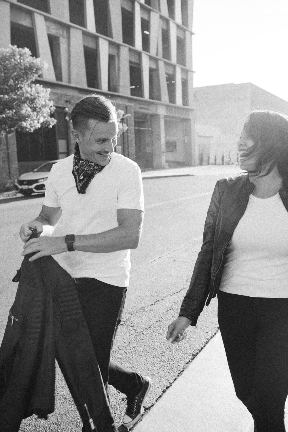Black and white photo of a smiling couple walking together on a city street, man in white tee, woman in leather jacket.