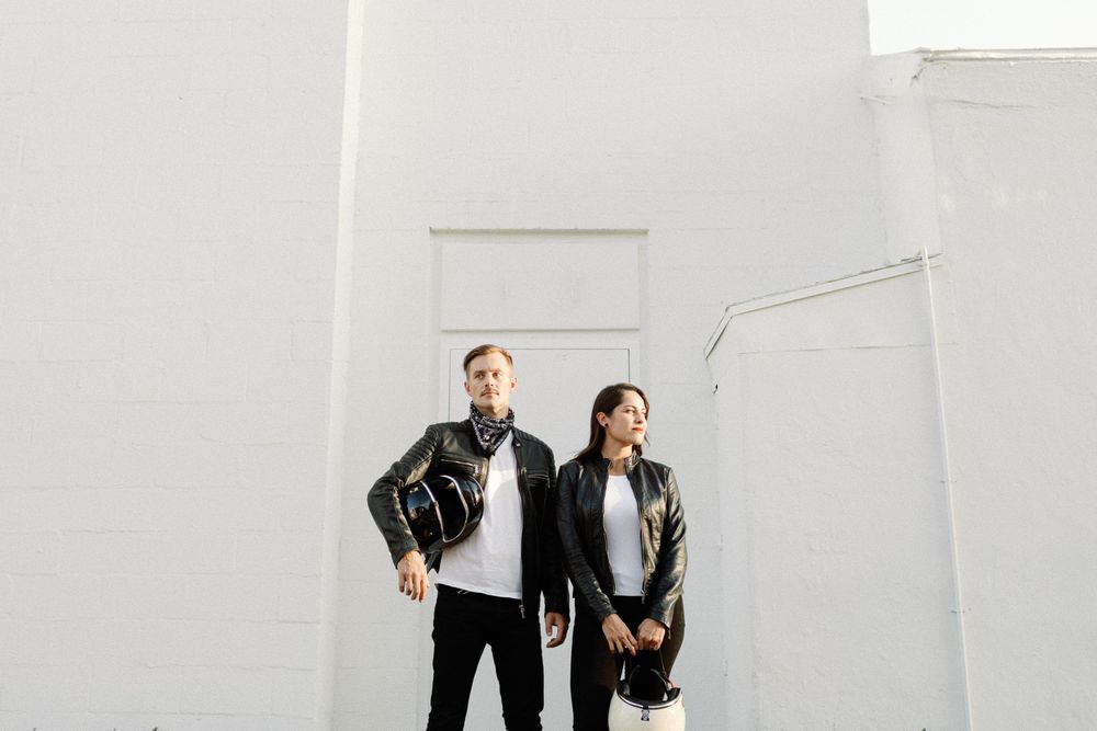 Couple in matching black leather jackets posing against a white wall outdoors.
