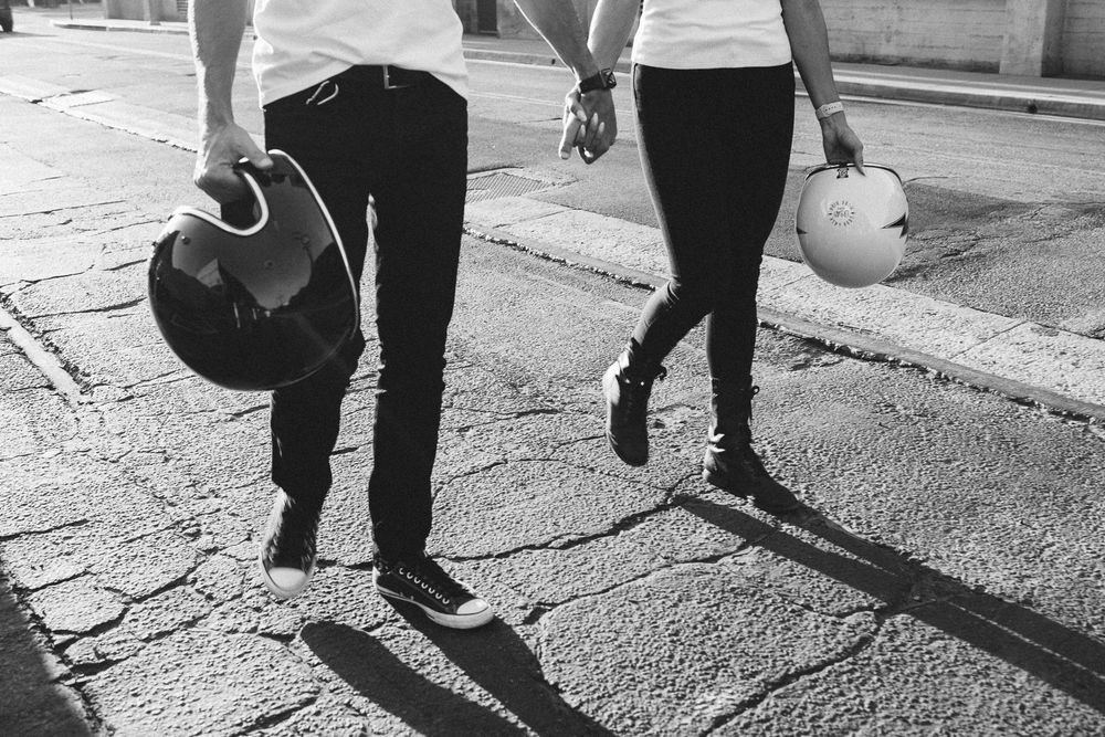 Couple walking hand in hand on a sunny street carrying motorcycle helmets in black and white.