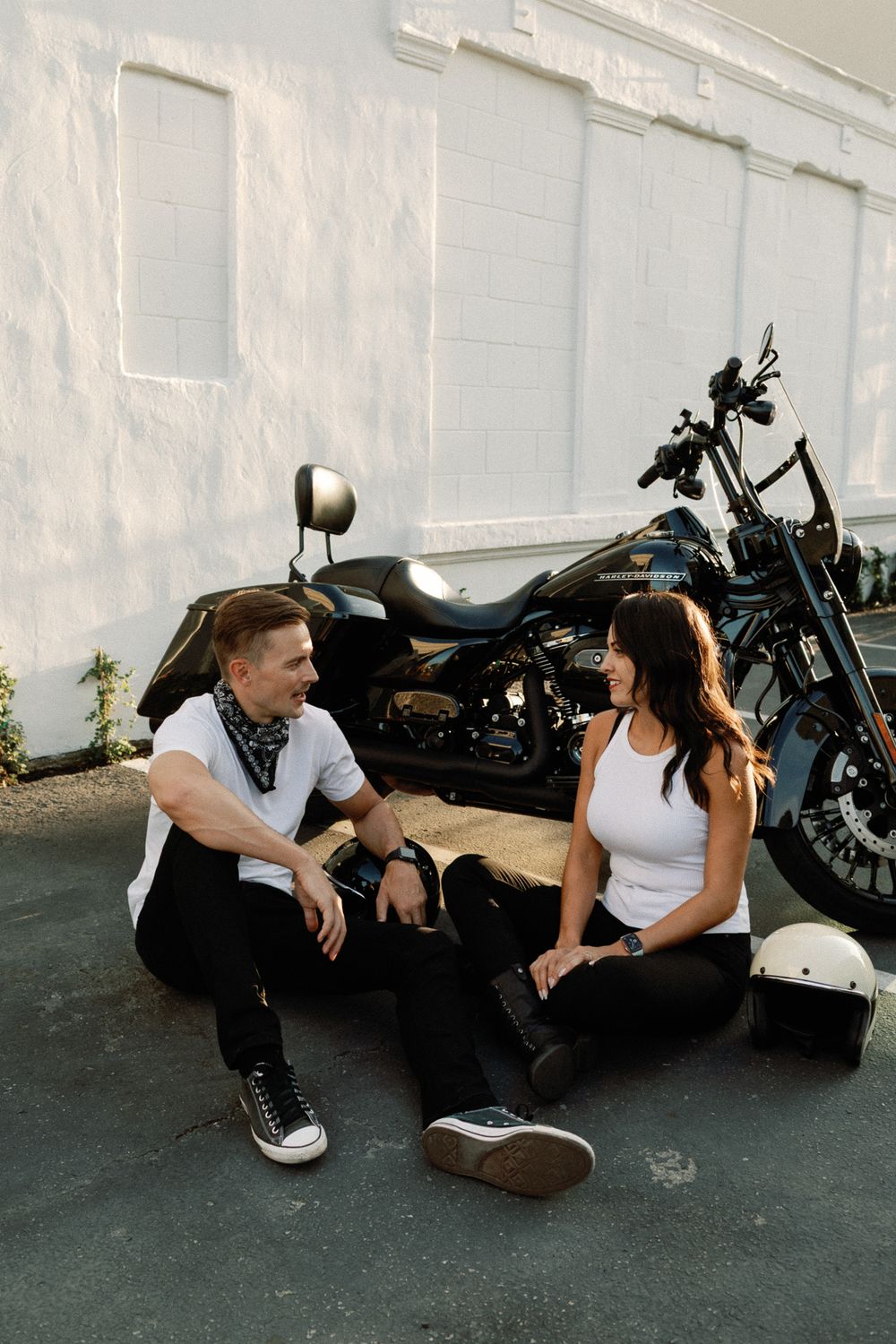 Smiling couple sitting together on pavement in front of a black Harley-Davidson motorcycle against a white wall.