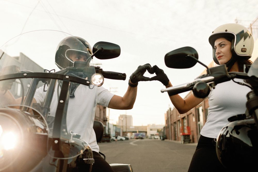 Two motorcyclists wearing helmets and gloves make a heart shape with their hands while sitting on their bikes.