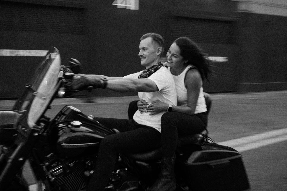Couple riding a Harley-Davidson motorcycle together on a city street in black and white photo.