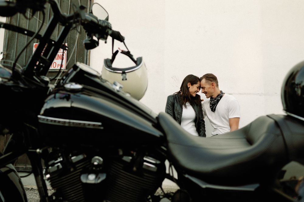 A romantic couple shares a tender moment together seated behind a black Harley-Davidson motorcycle.